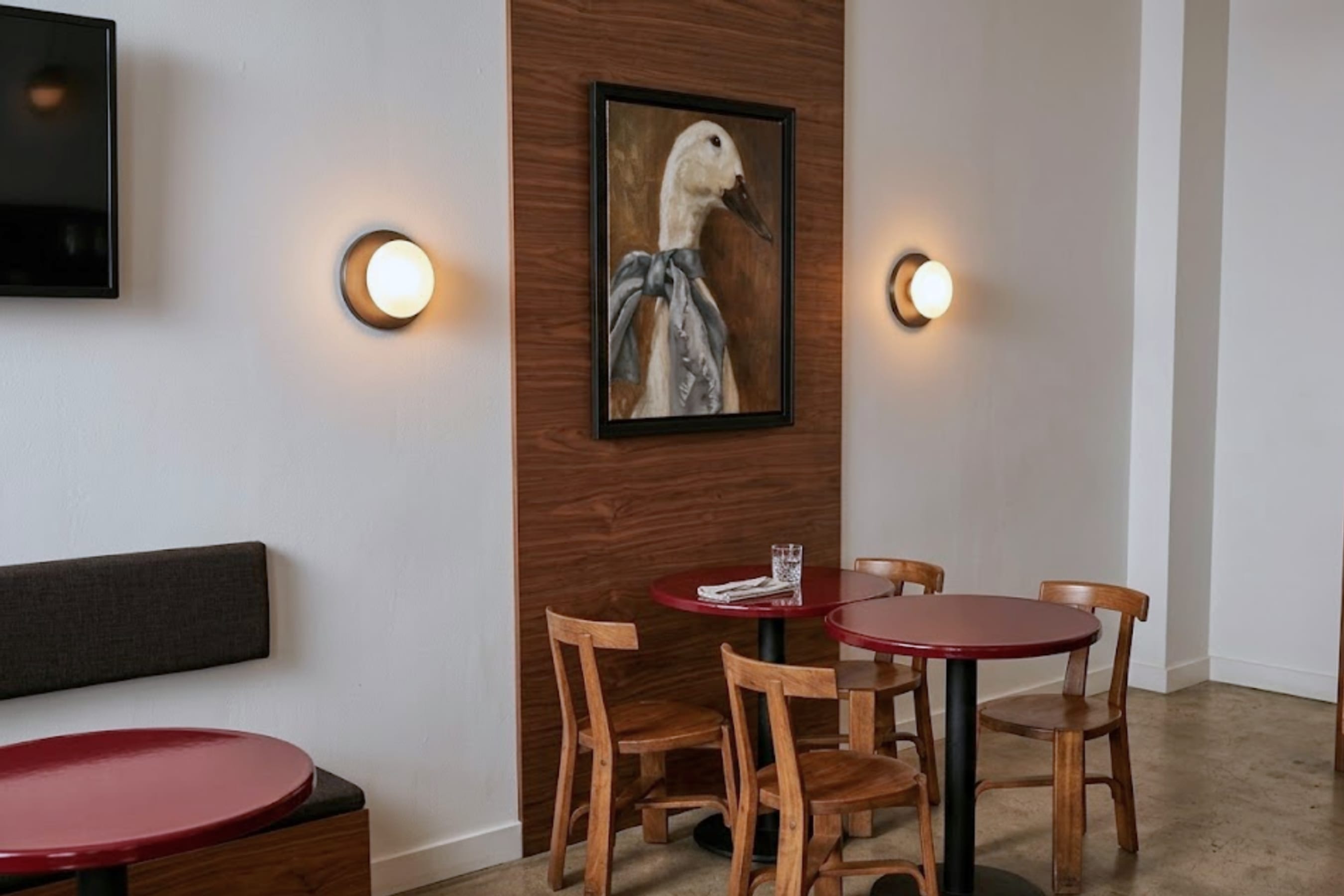 Seating nook with round red tables, two wall lights and a framed painting on a timber panel.