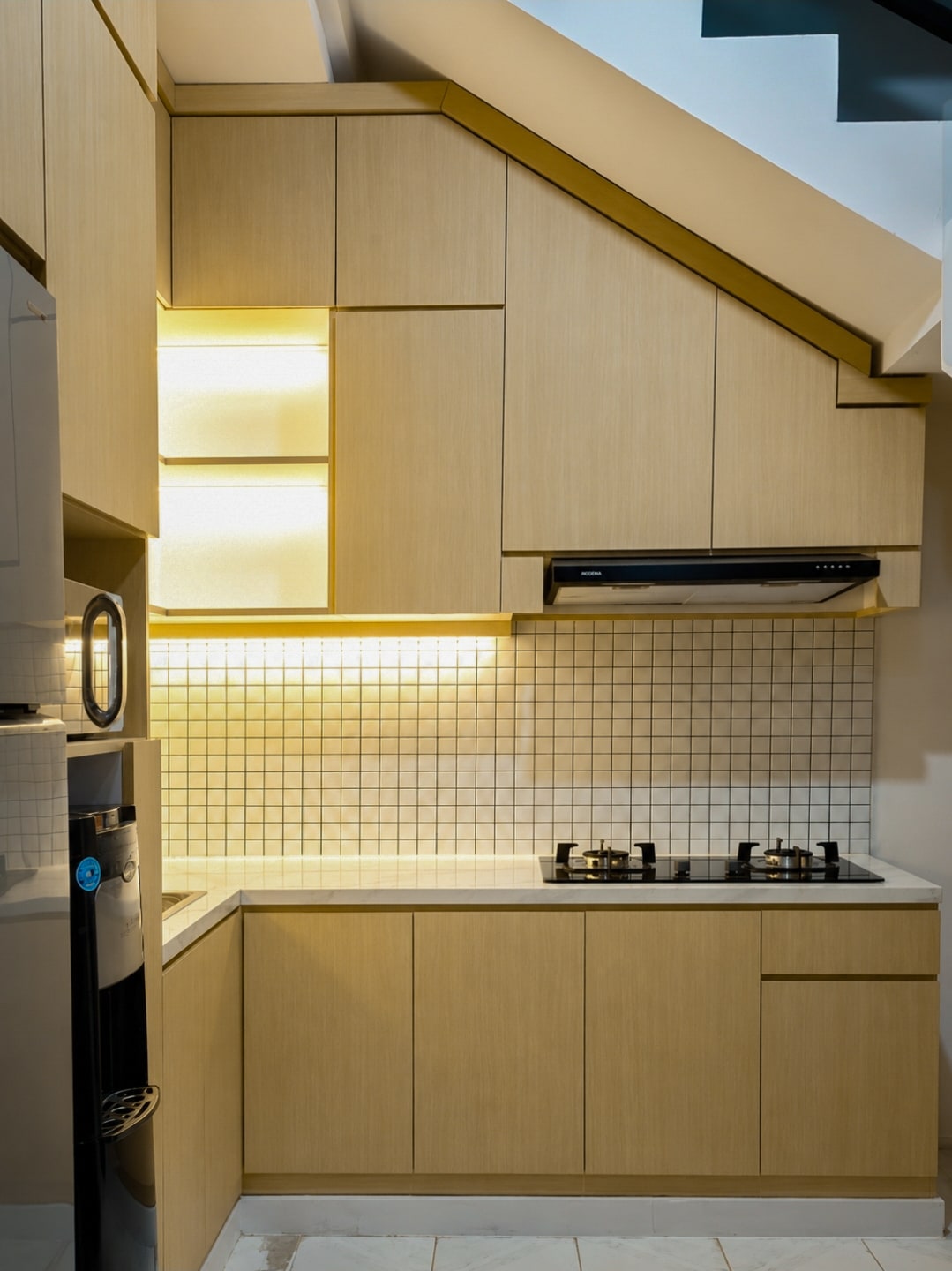 A light timber-grain kitchen fitted beneath a staircase, with lit open niches.
